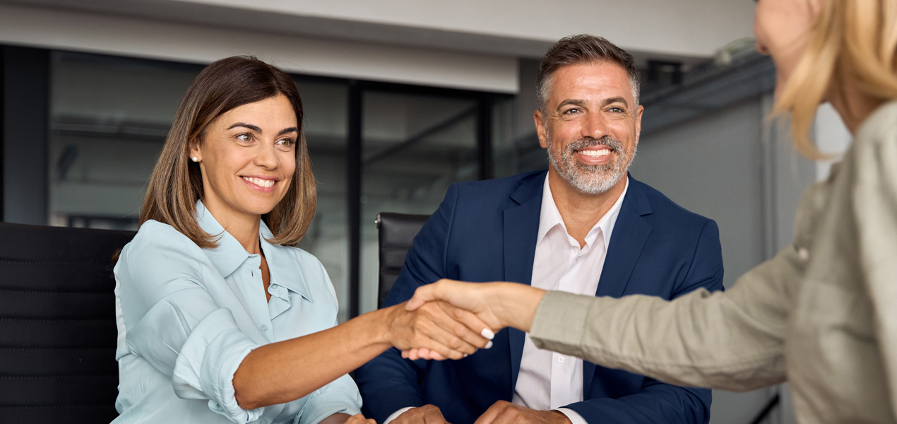 Two professionals, a woman in a light blue blouse and a man in a blue suit, smile as they shake hands with a person in the foreground. They are seated in an office setting.