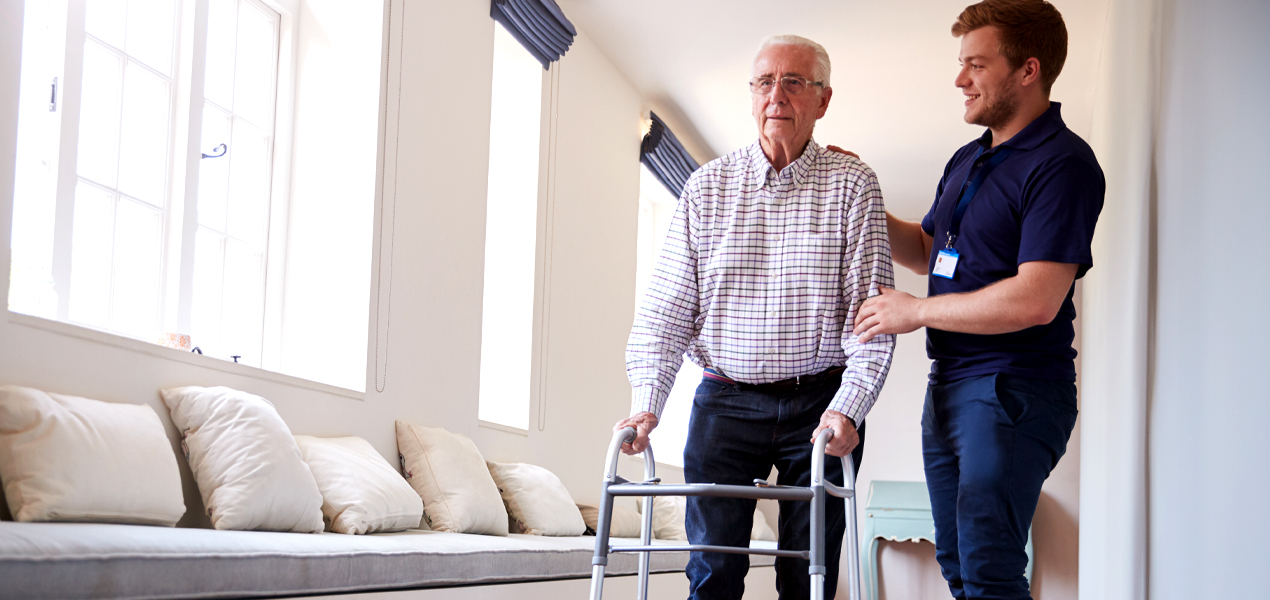 An elderly man using a walker is assisted by a young male caregiver in a well-lit hallway with bench seating and cushions. The caregiver smiles, offering support as they move together.