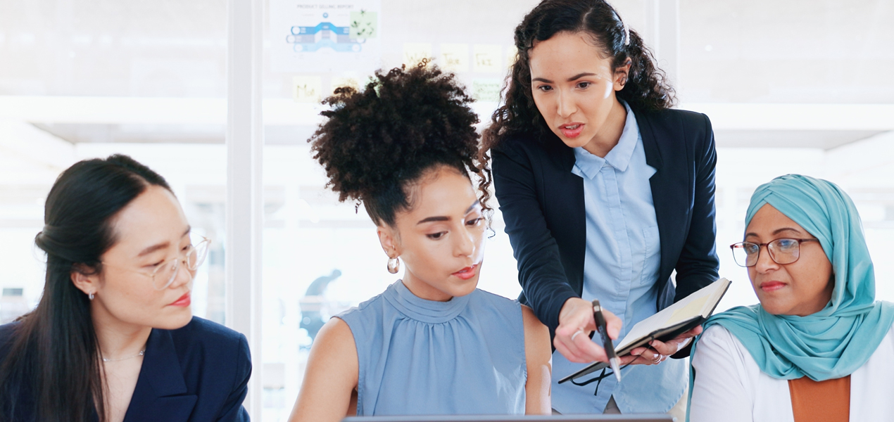 A group of women in business attire collaboratively reviewing information on a laptop.A group of women in business attire collaboratively reviewing information on a laptop.